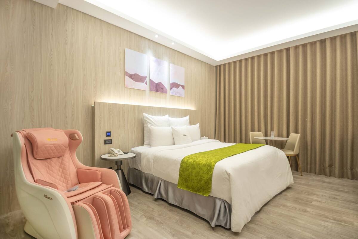 A hotel room with a bed, a massage chair, a small table, and a chair.