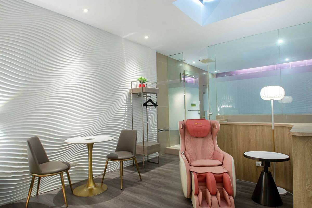 A modern room with a unique textured wall, a massage chair, a small table, and a glass shower area.