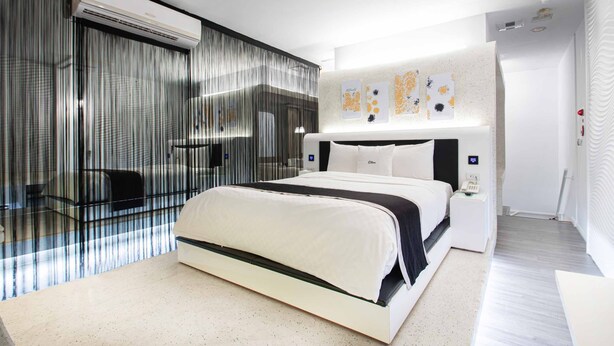 A modern bedroom with a glass partition, a bed with white bedding, a bedside table, and a wall-mounted light fixture. - 168 Motel-Taoyuan (Taoyuan City)