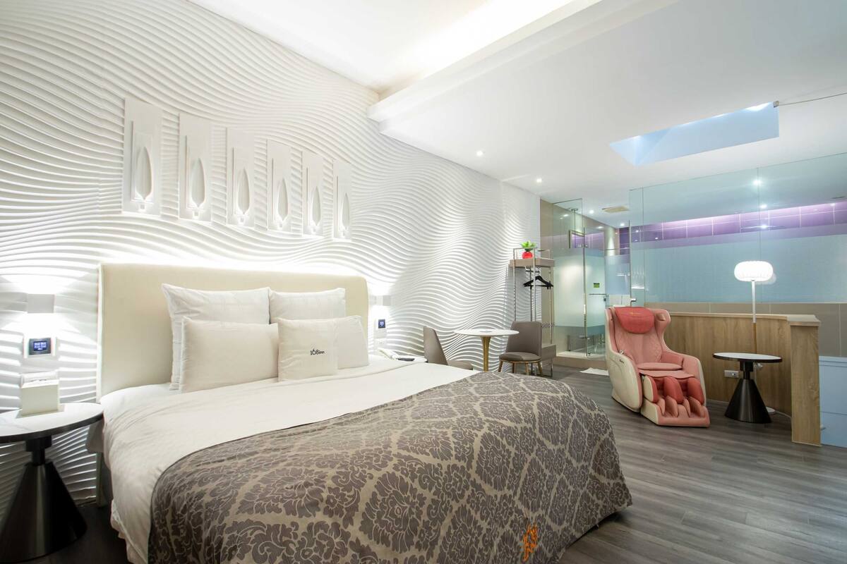 A modern hotel room with a large bed, a massage chair, a small table, and a glass partition wall.