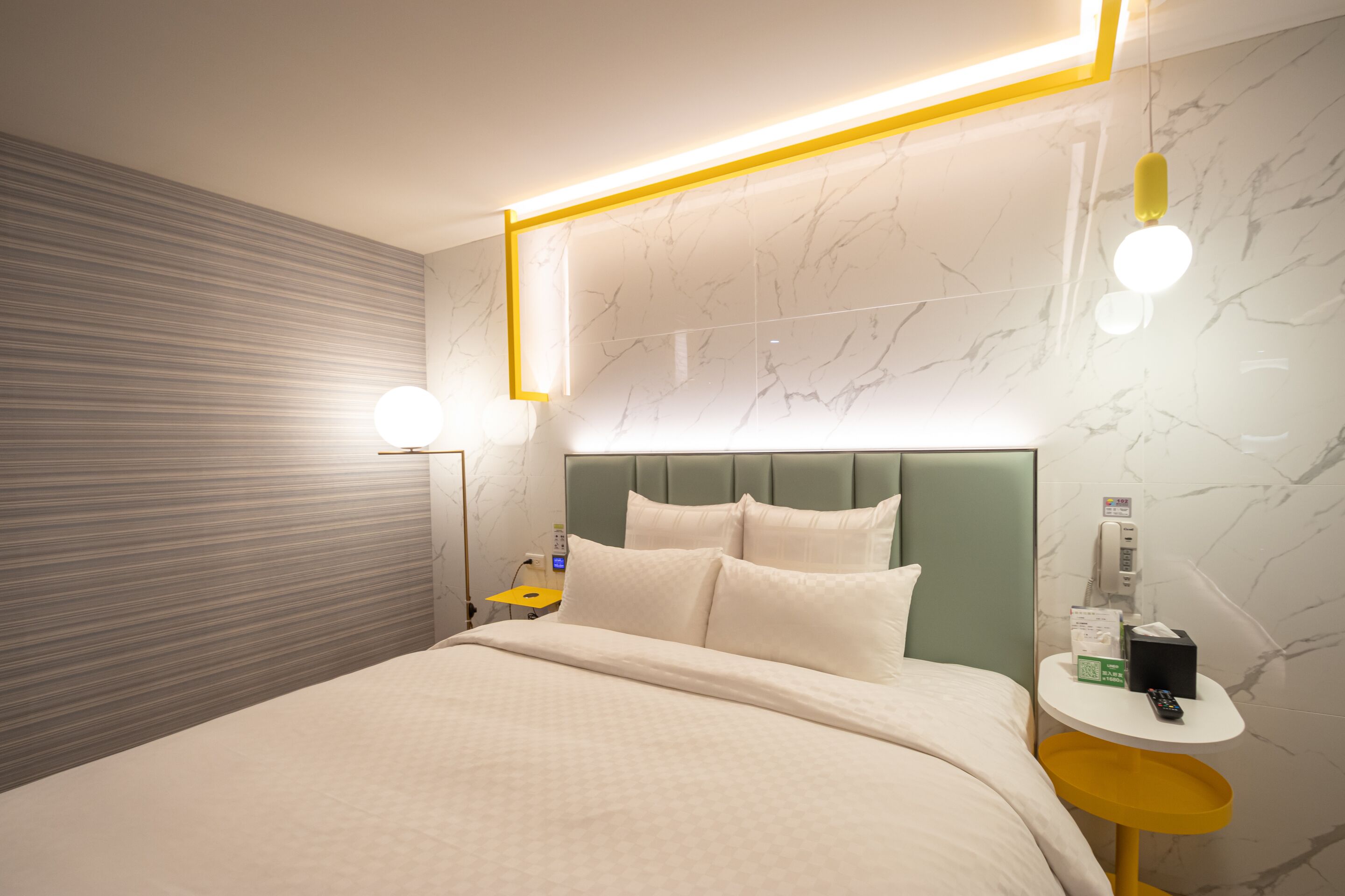 A modern hotel room with a neatly made bed, a bedside table with a lamp, and a wall-mounted phone.