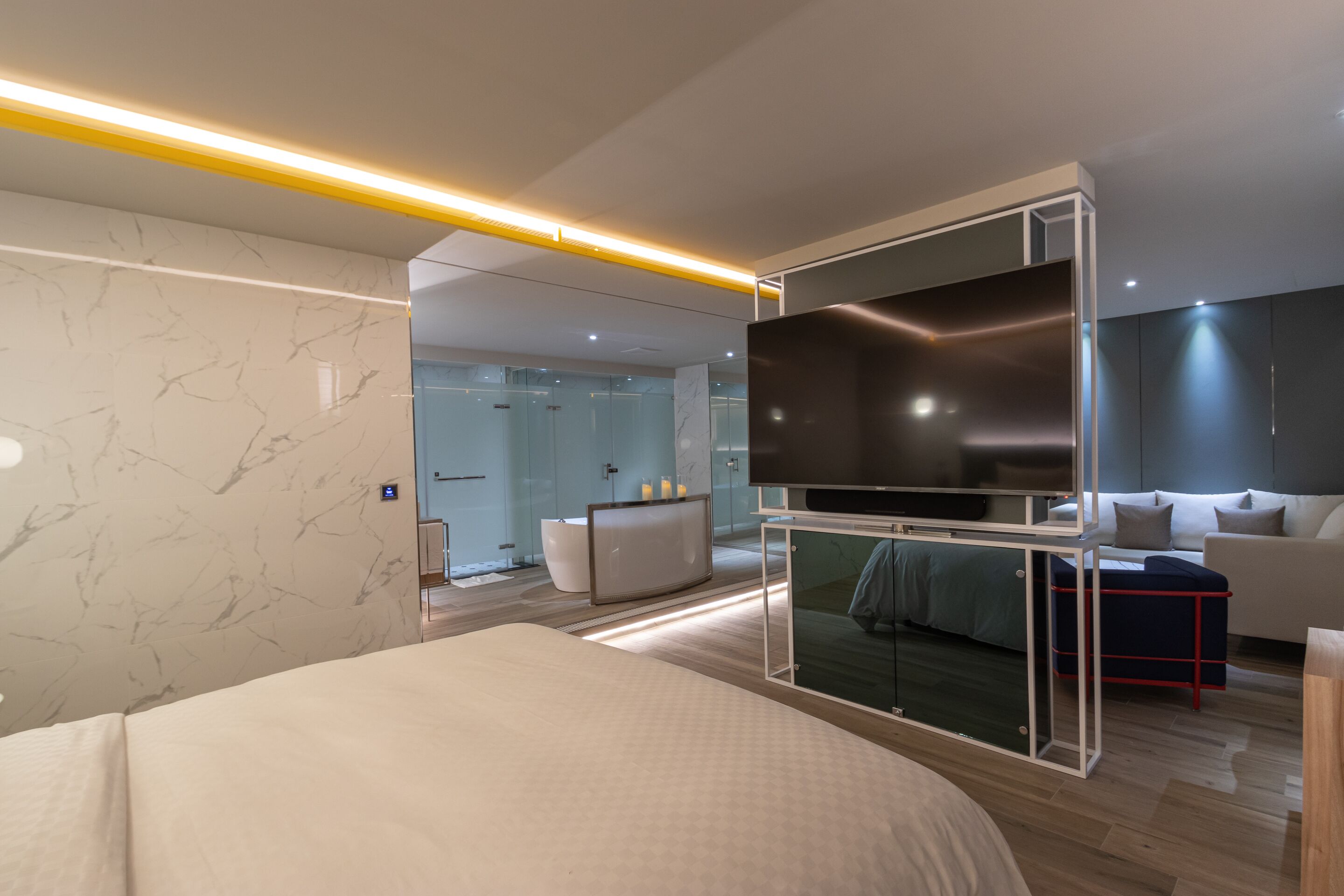A modern hotel room with a large bed, a TV, a glass partition, a sofa, and a bathroom visible in the background.