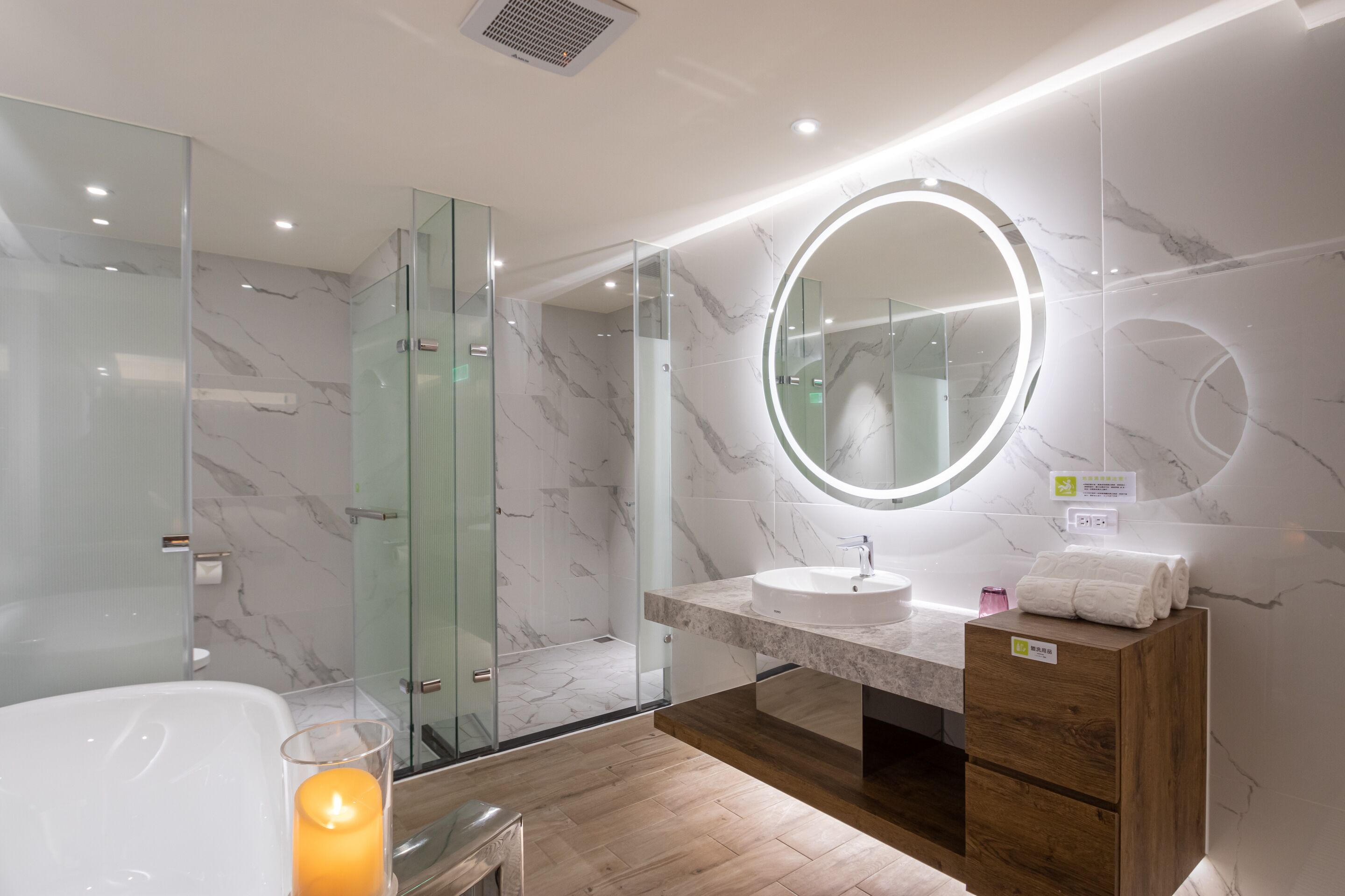Un baño moderno con ducha de vidrio, un espejo redondo con luz, tocador de madera y una vela encendida.