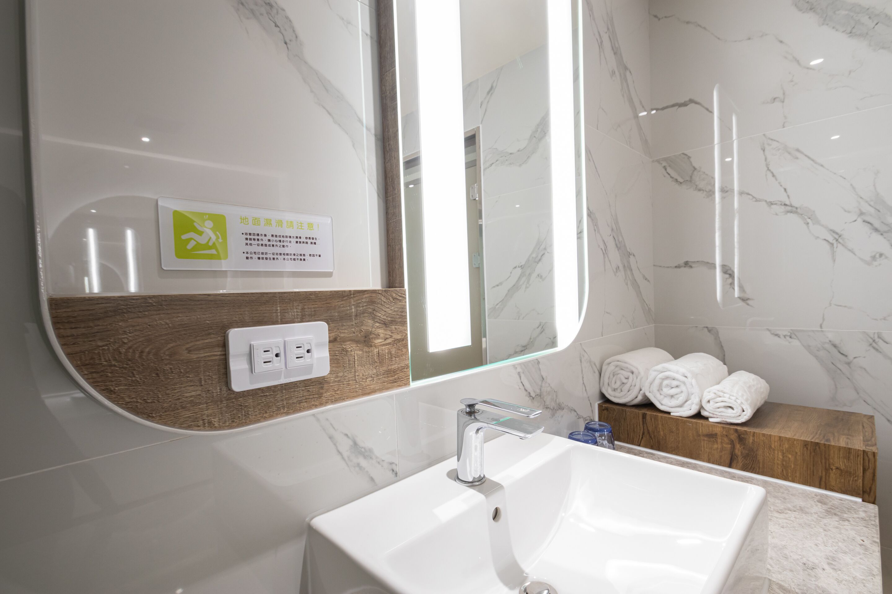 A modern bathroom with a white sink, a mirror with a wooden frame, and a marble wall.