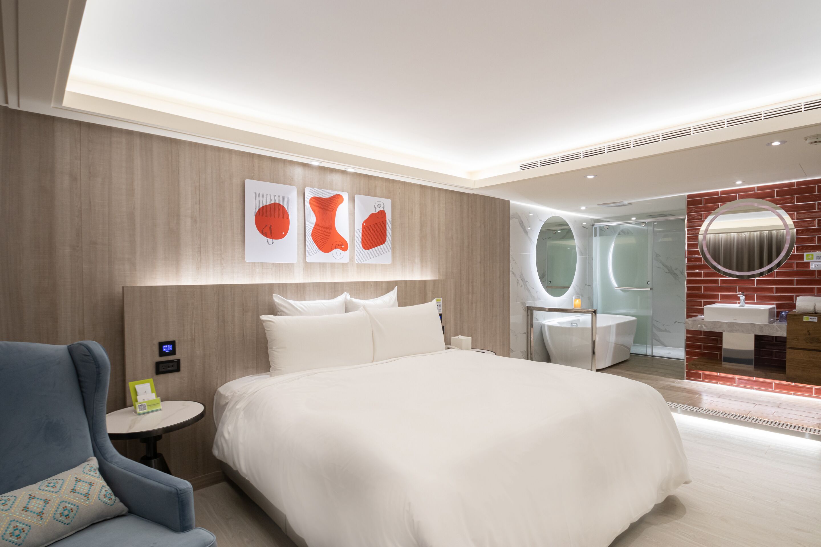 Una habitación de hotel moderna con una cama grande, un espejo redondo, una mesita pequeña y un área de descanso con una silla.