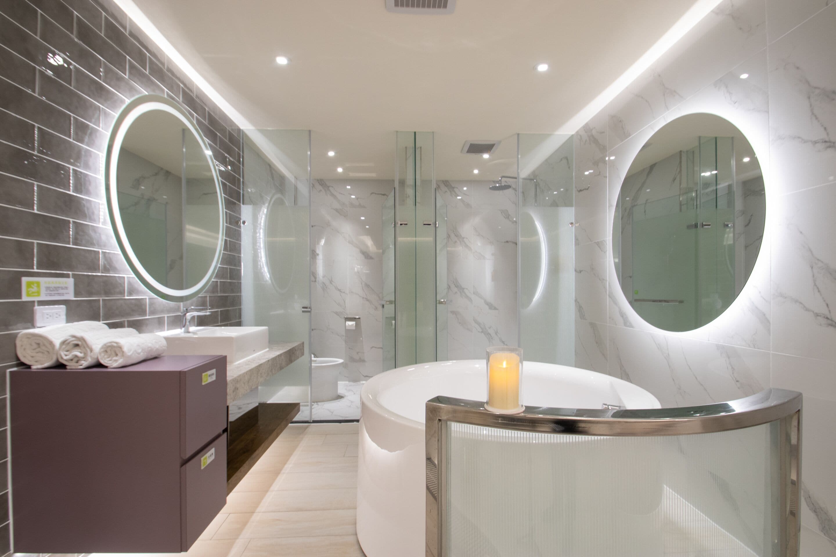 Un baño moderno con pared de mármol, un espejo redondo grande, una bañera y un box de ducha con mampara de vidrio.