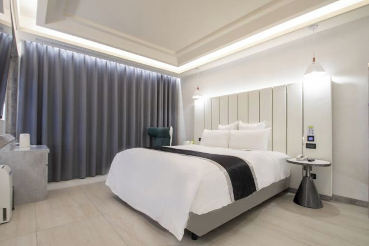 Una habitación de hotel moderna con una cama grande, una mesita de noche, una lámpara y un ventanal con cortinas.