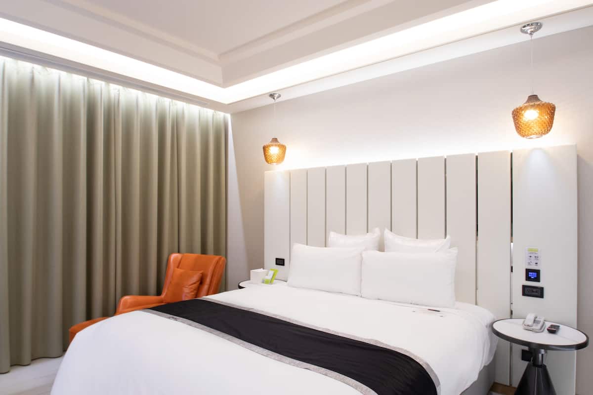 Una habitación de hotel moderna con una cama grande, una mesita de noche, una silla y un ventanal con cortinas.