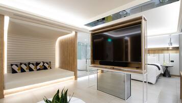A modern living room with a large flat-screen TV mounted on a wooden frame, a white sofa with patterned cushions, a glass coffee table with a plant, and a minimalist design.