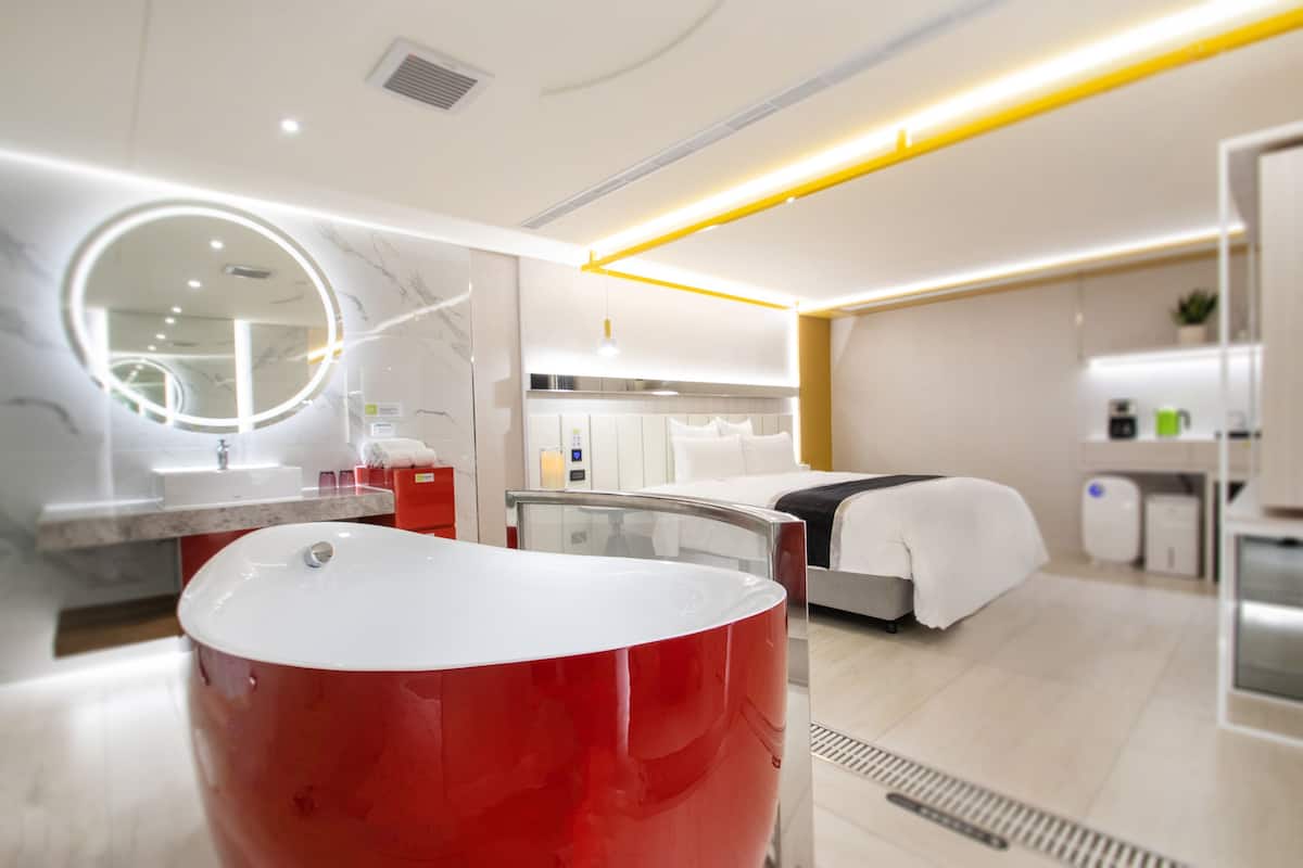 Una habitación de hotel moderna con una bañera roja, un espejo redondo, una cama, una mesita de noche y una pequeña kitchenette.