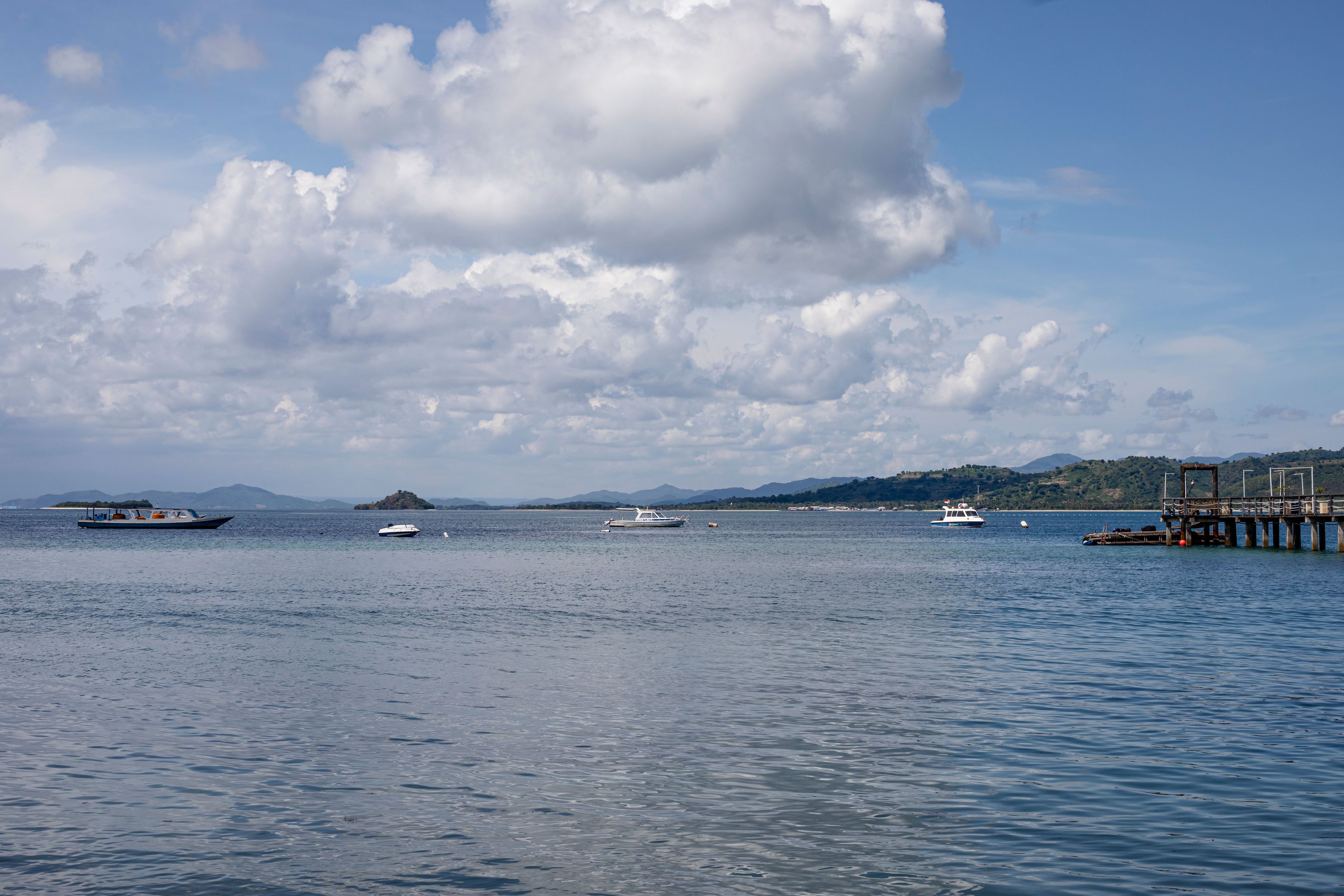Un cuerpo de agua tranquilo con barcos, un muelle y montañas distantes bajo un cielo parcialmente nublado.