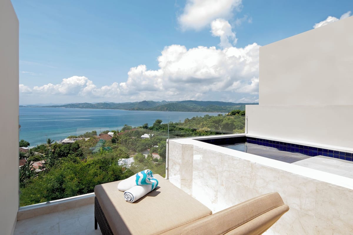 A balcony with a view of the sea and hills, a small pool, and a lounge chair with a towel.