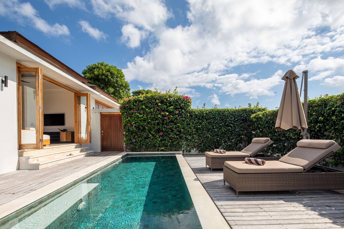 A modern outdoor pool area with a wooden deck, lounge chairs, and a hedge.