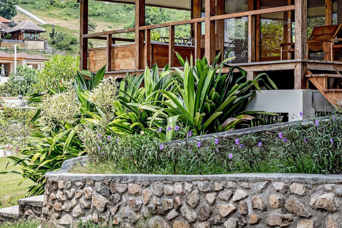 A wooden building with a covered patio area, surrounded by lush greenery and a stone wall.