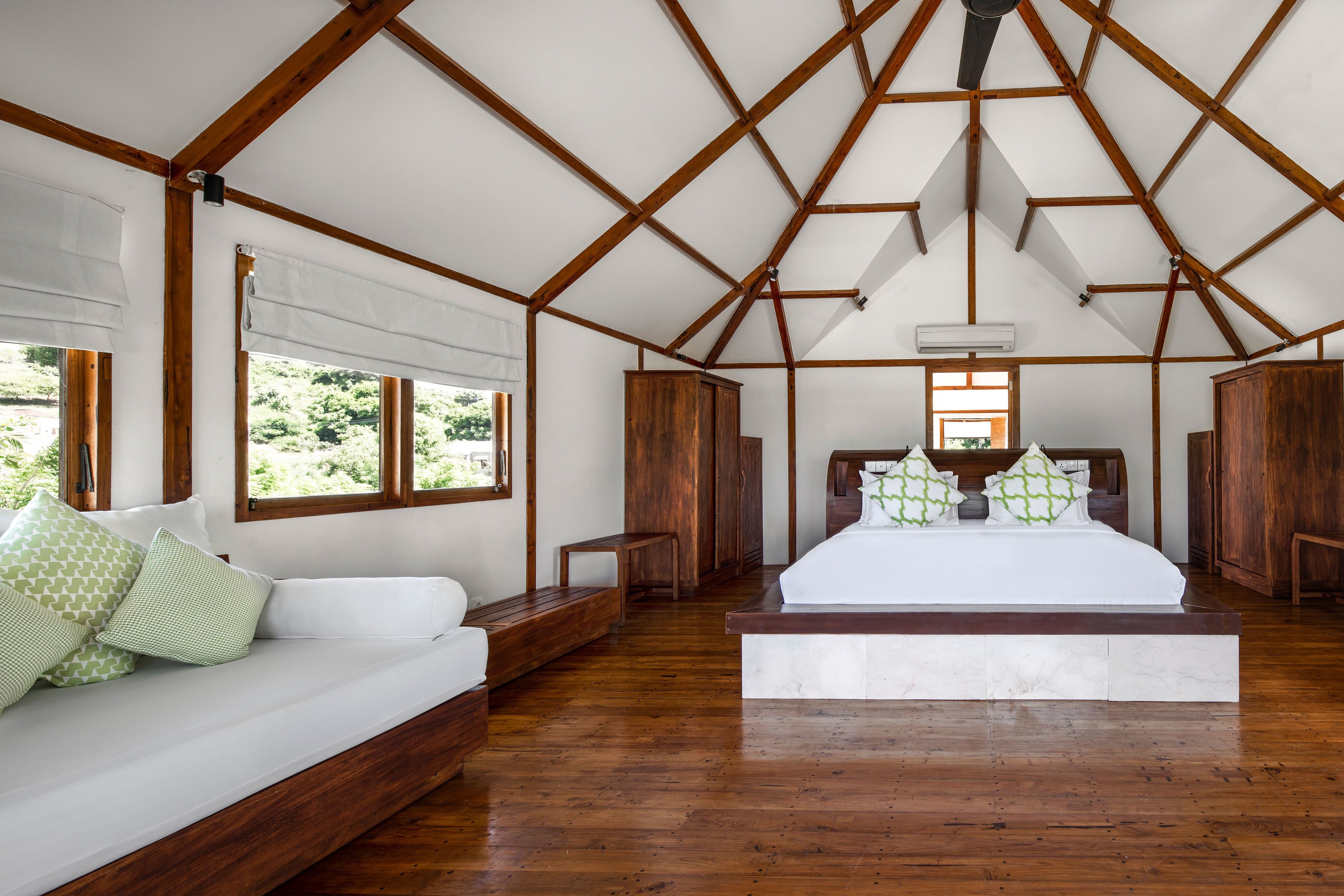 Een slaapkamer met een houten plafond, een groot bed, een bank en een raam met uitzicht op groen.