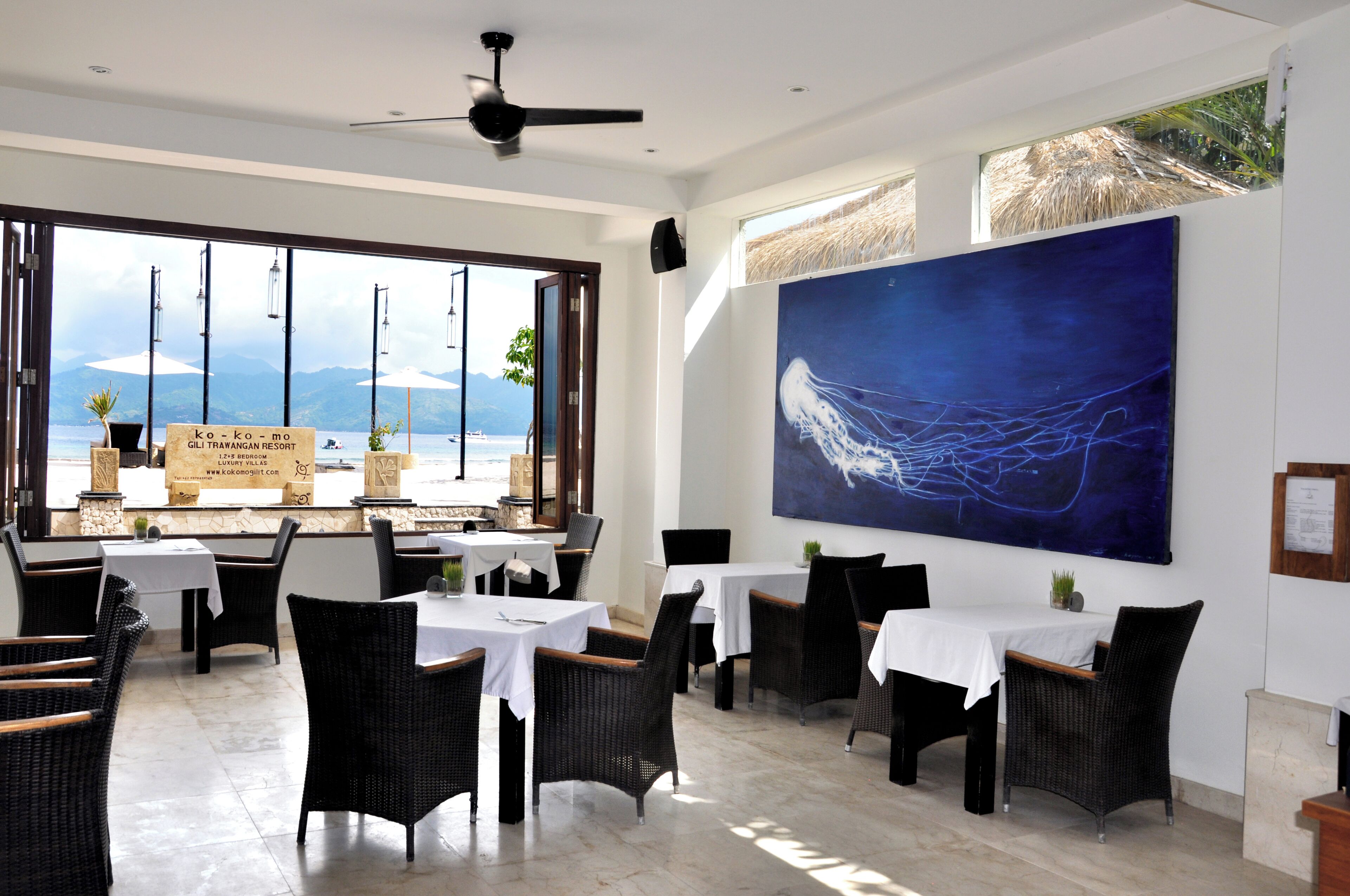 A restaurant with tables and chairs, a large painting on the wall, and a view of the sea.