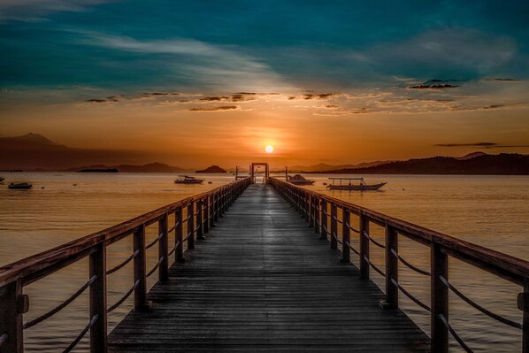 A wooden pier leading to a sunset over calm water with boats and distant mountains. - Kokomo Resort Gili Gede (Gili Gede)