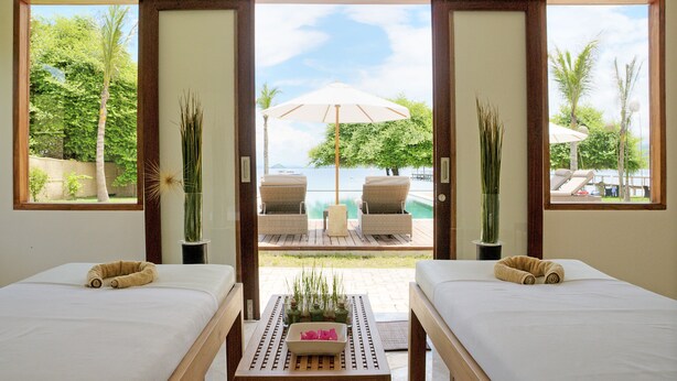 A hotel room with two beds, a small table with a vase, and a view of a pool and palm trees. - Kokomo Resort Gili Gede (Gili Gede)