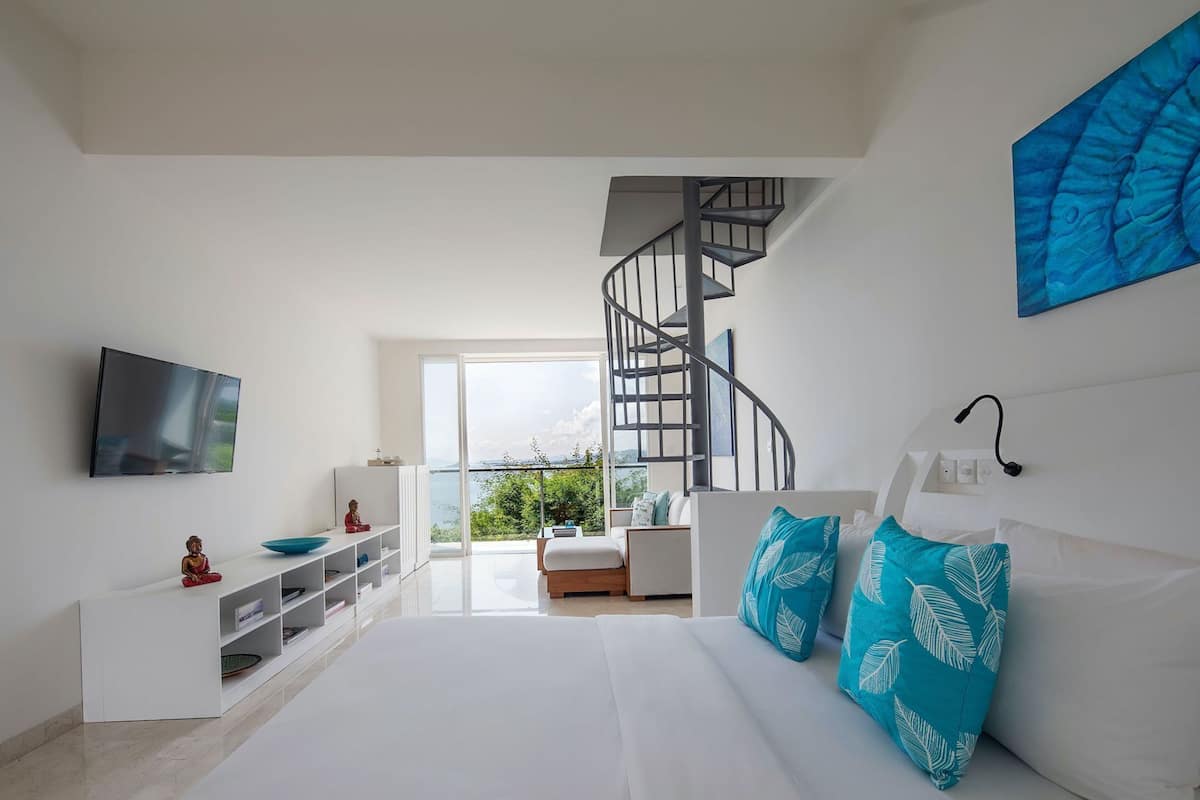 A modern bedroom with a white bed, a TV, a staircase, and a view of the outdoors.