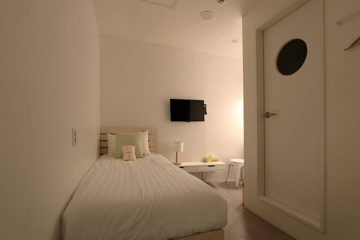 Ein Hotelzimmer mit Bett, Schreibtisch, Sessel, Wand-Fernseher und einer Tür mit rundem Fenster.