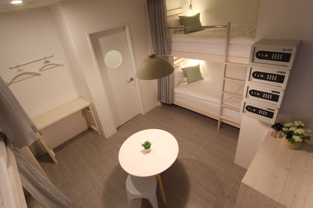 Ein kleines Zimmer mit Etagenbetten, einem Tisch mit Pflanze und einem an der Wand befestigten Bedienfeld.