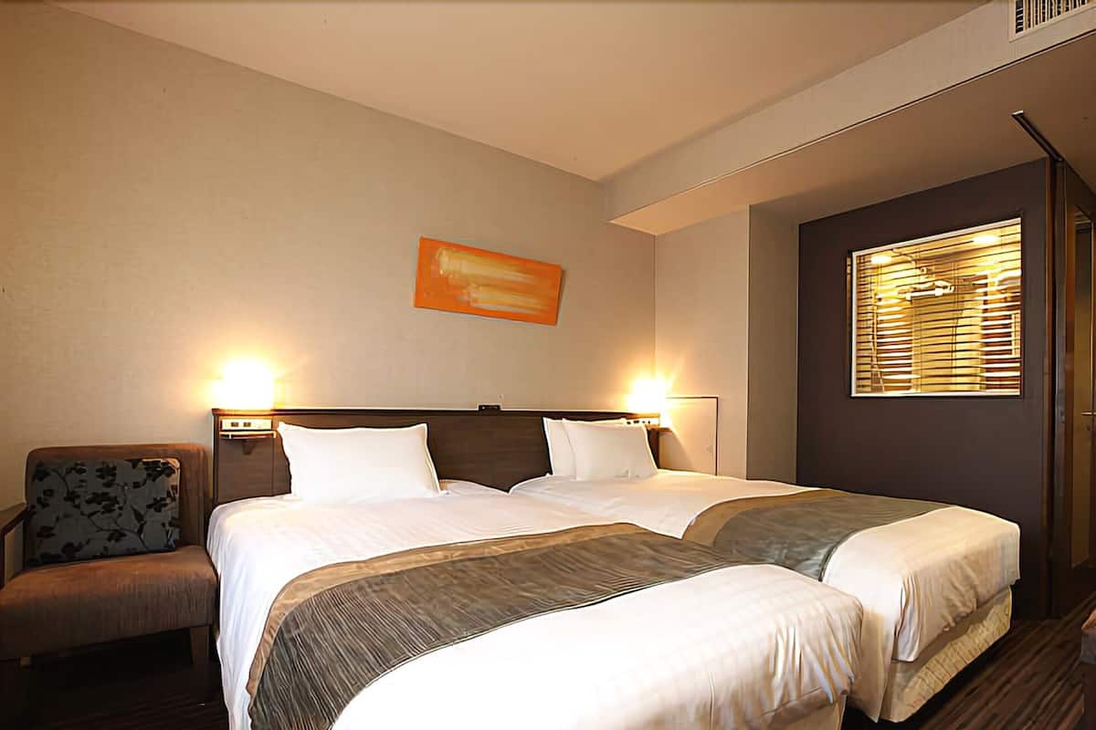 Une chambre d’hôtel avec deux lits, une chaise et une fenêtre avec des stores.