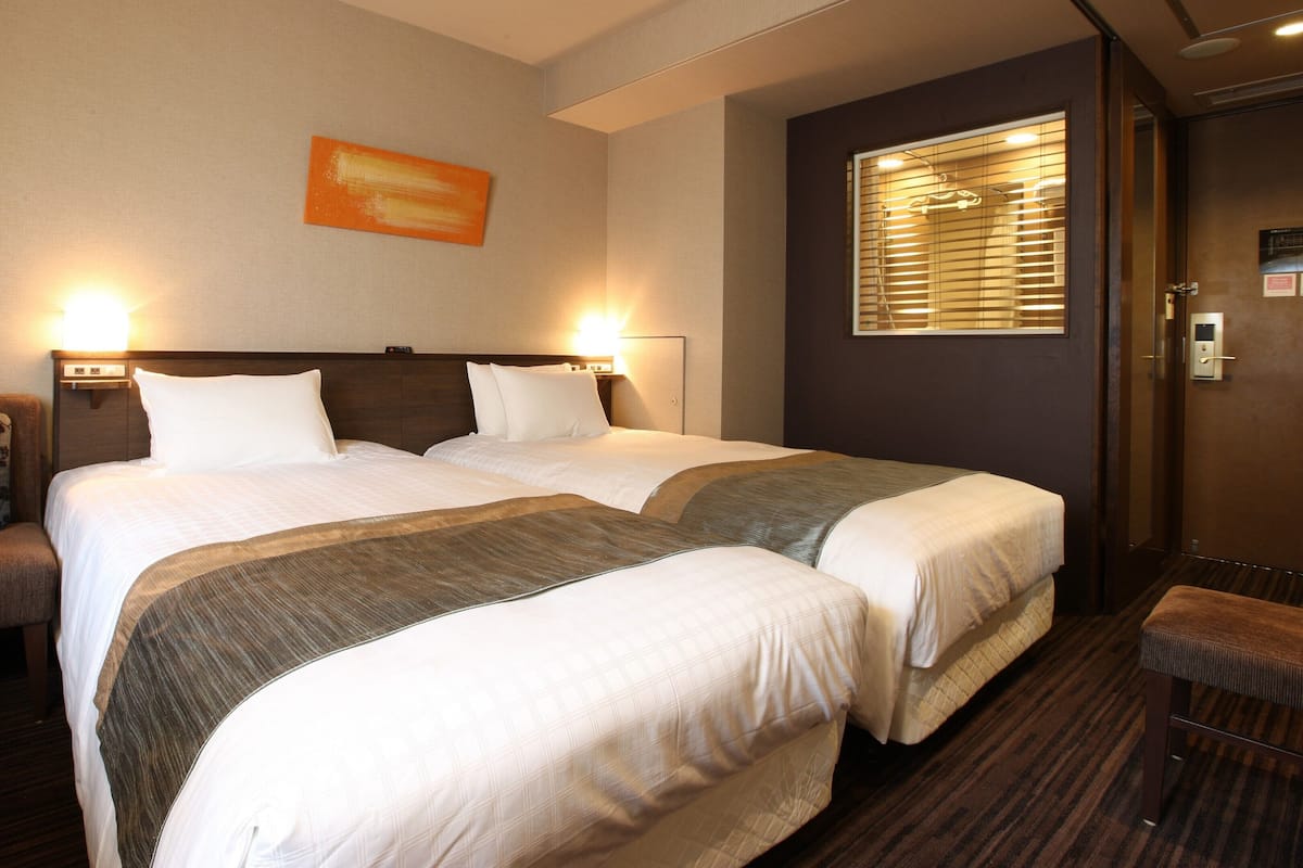 Une chambre d’hôtel avec deux lits, une chaise, une fenêtre avec des stores et une porte avec une serrure.