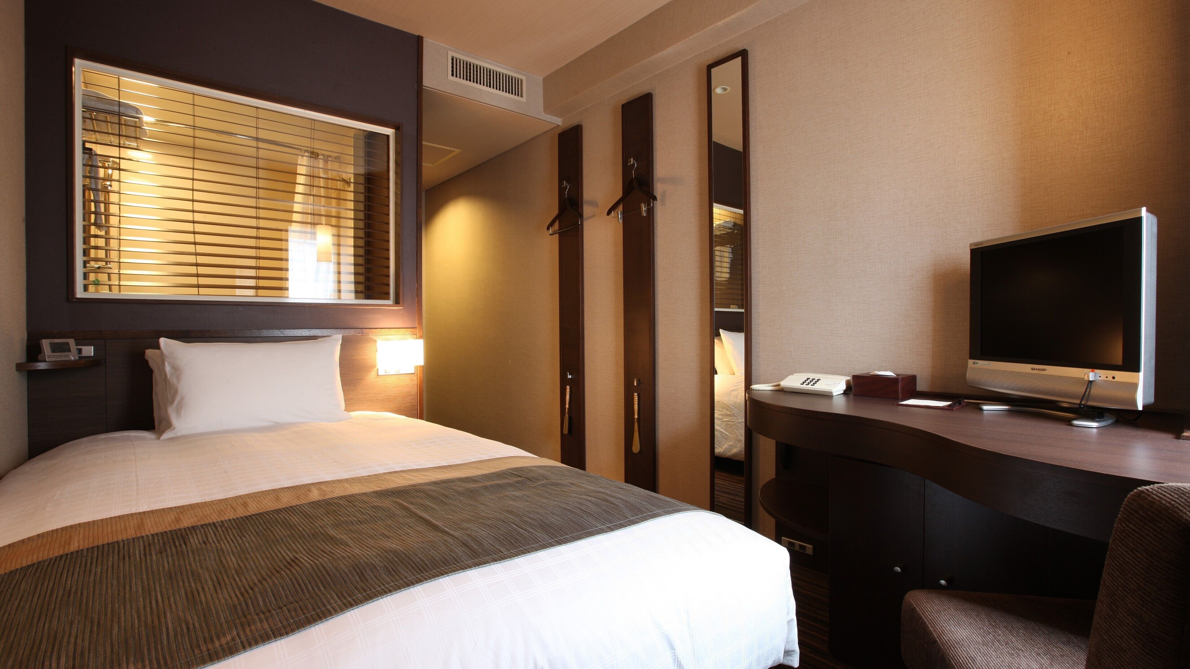 A hotel room with a bed, bedside lamp, desk with a TV, and a mirror.
