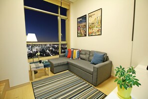 A modern living room with a grey sectional sofa, a striped rug, a small side table with a lamp, and a cityscape view through large windows. - The Sweet Castle Apartment (Seoul)