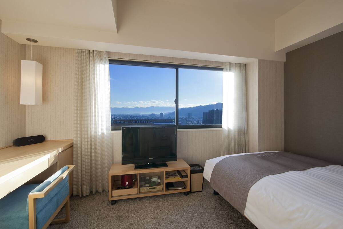 A hotel room with a bed, a desk with a television, and a view of a cityscape through a large window.