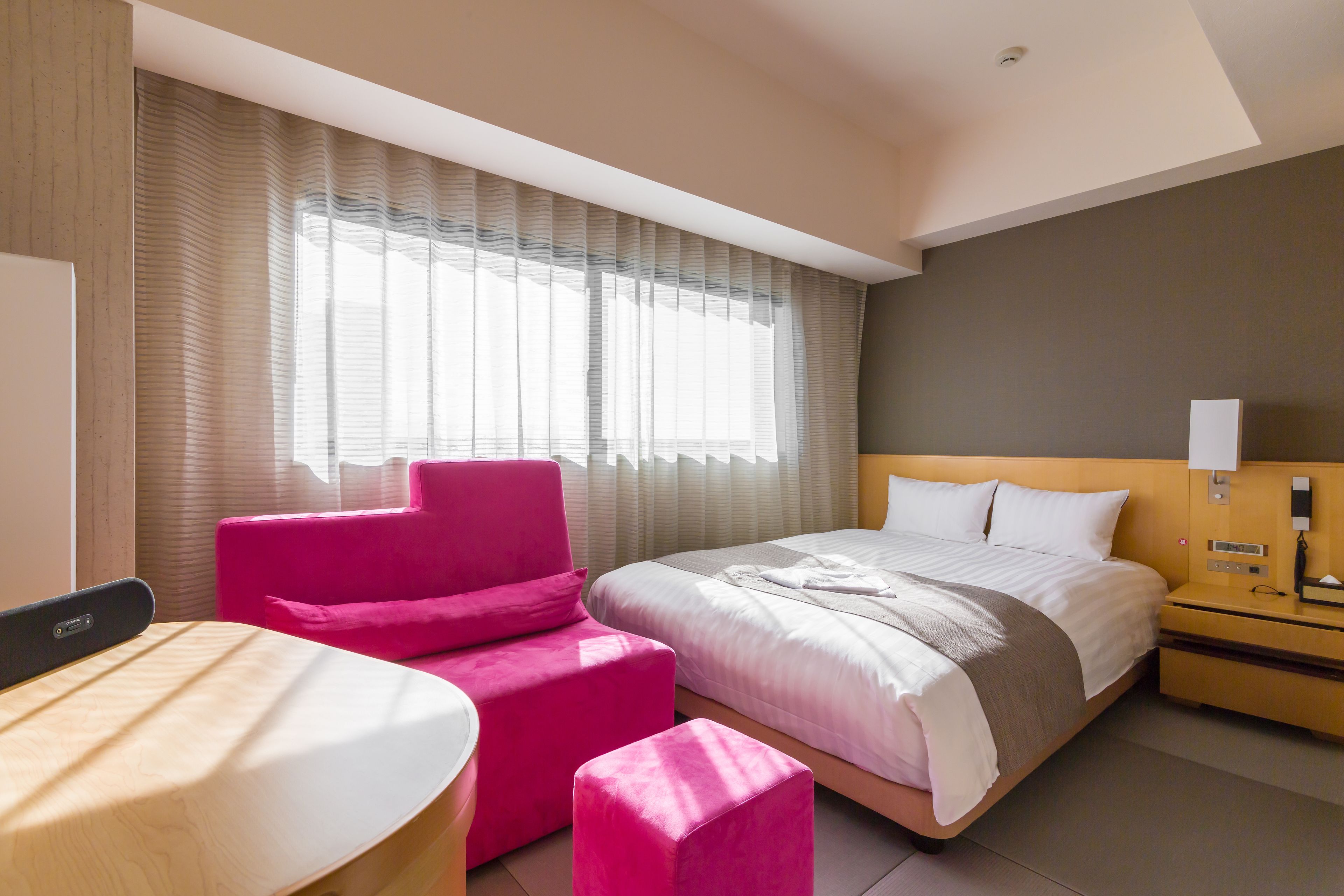 A modern hotel room with a bed, a pink armchair, a wooden desk, and a large window with curtains.