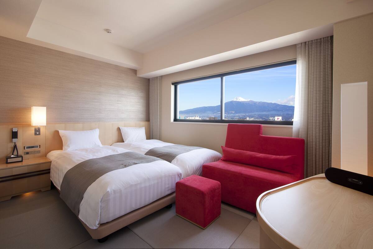 A hotel room with two beds, a red chair, a desk, and a large window with a mountain view.