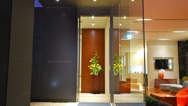 A hotel entrance with a glass door, a reception desk, and a seating area with orange chairs. - Cocochee Hotel Numazu (Numazu)