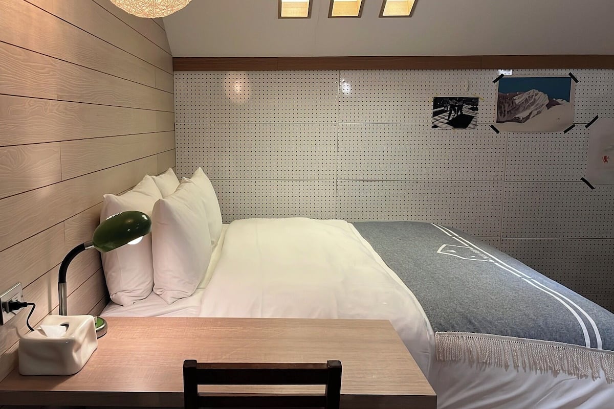 A hotel room with a wooden wall, a bed with white bedding, a desk with a lamp, and a wall-mounted shelf.