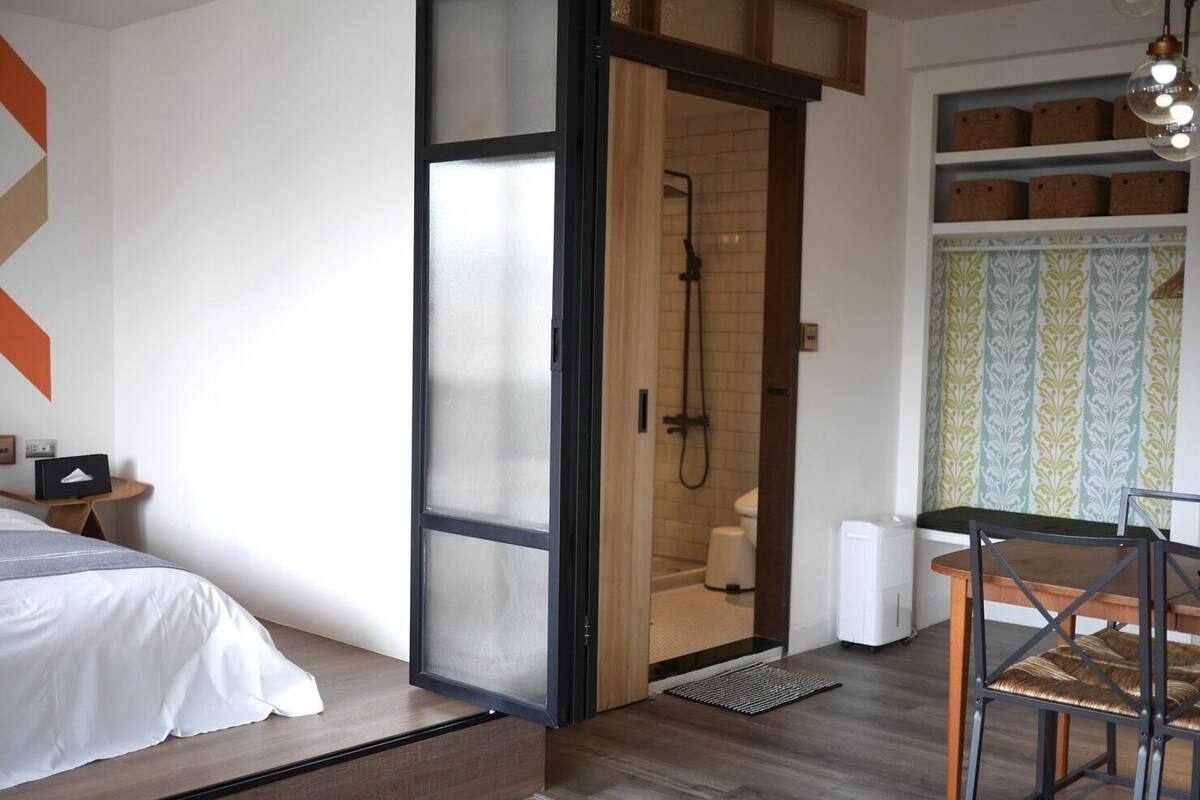 A modern bedroom with a bed, a wooden door leading to a shower, and a dining area with a table and chairs.