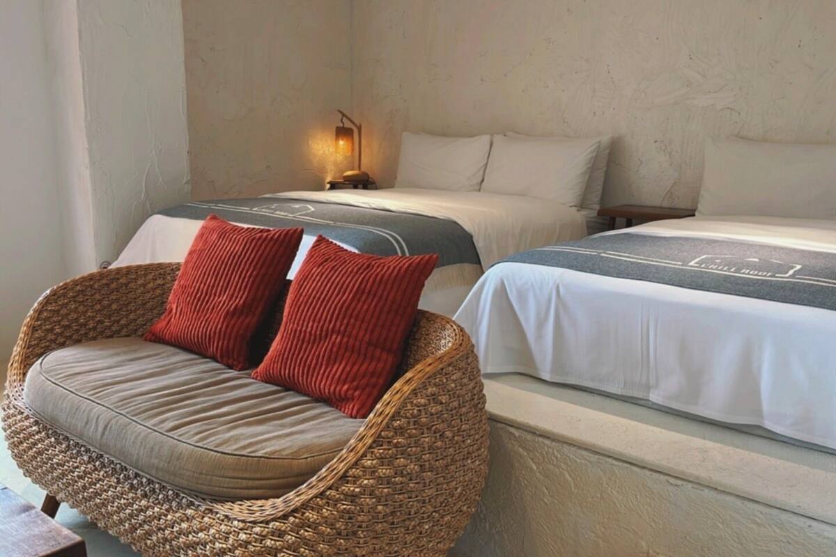 A room with a wicker chair with cushions, a bed with white bedding and a grey headboard, and a small table with a lamp.