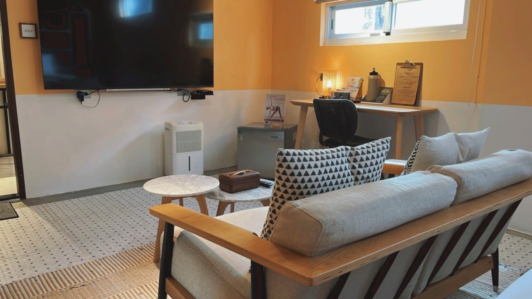 A modern room with a sofa, two small round tables, a desk with a chair, a television, and a window.