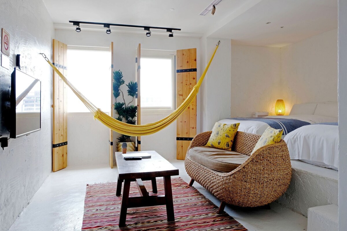 A room with a hammock, a bed, a table, and a chair.
