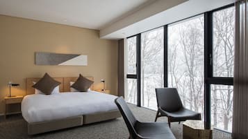 A modern hotel room with a large bed, two chairs, a nightstand, and a large window offering a view of trees.