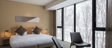 A modern hotel room with a large bed, two chairs, a nightstand, and a large window offering a view of trees.