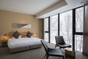Habitación de hotel moderna con una cama grande, dos sillas, una mesita de noche y un amplio ventanal con vistas a árboles.