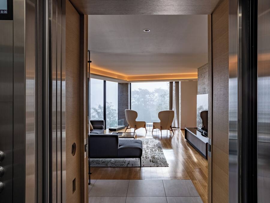 An interior view of a modern elevator leading to a living area with a sofa, chairs, and a large window.