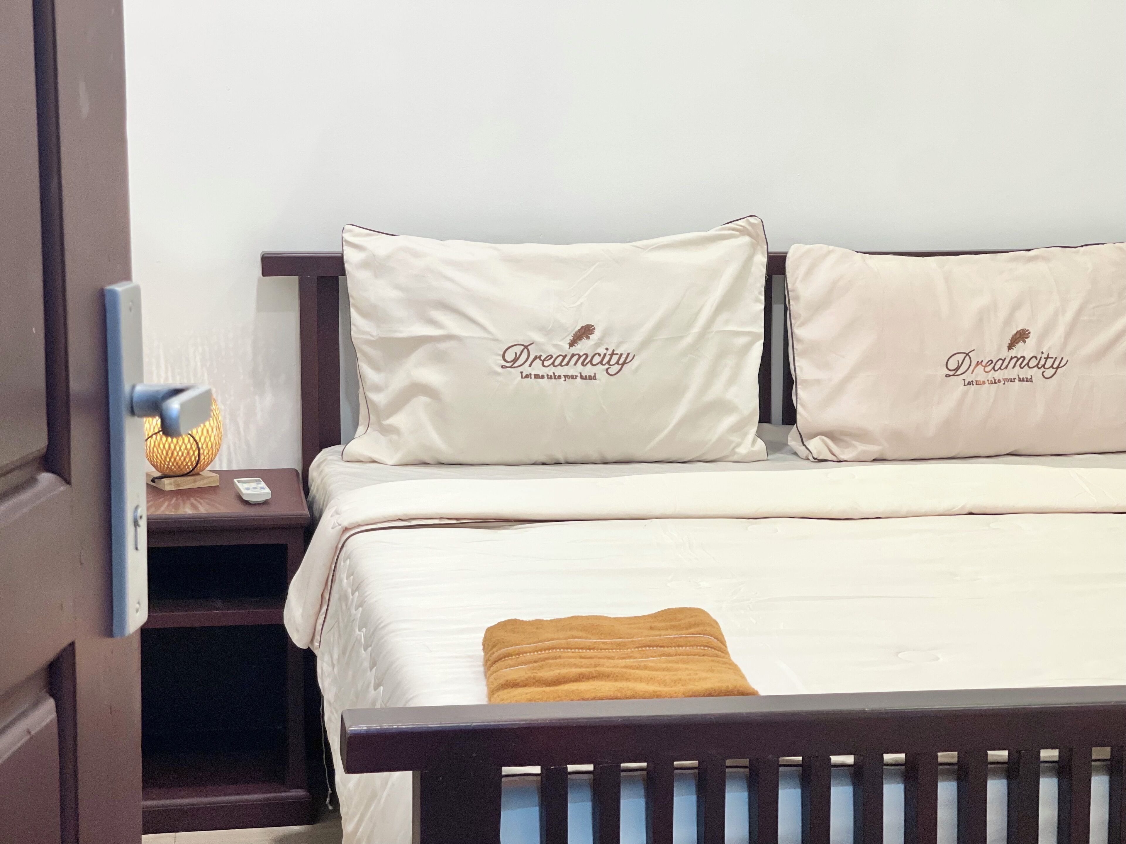 A neatly made bed with two pillows labeled "Dreamcity" and a bedside table with a lamp and a remote control.