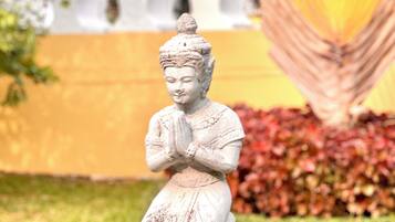 A statue of a figure with hands in a prayer position, set on a pedestal in a garden with a yellow wall and red foliage in the background.