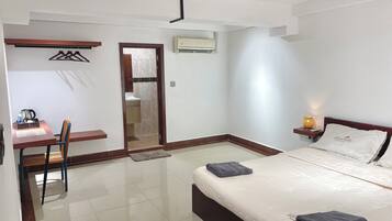 A hotel room with a bed, a desk with a chair, a small table with a lamp, and an air conditioning unit.