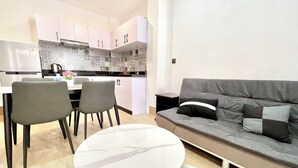 A modern kitchen with white cabinets, a black and white patterned backsplash, and a stainless steel refrigerator. There is a dining area with a white table and two chairs. A grey sofa with black and white cushions is against the wall. - RS guesthouse (Phnom Penh)