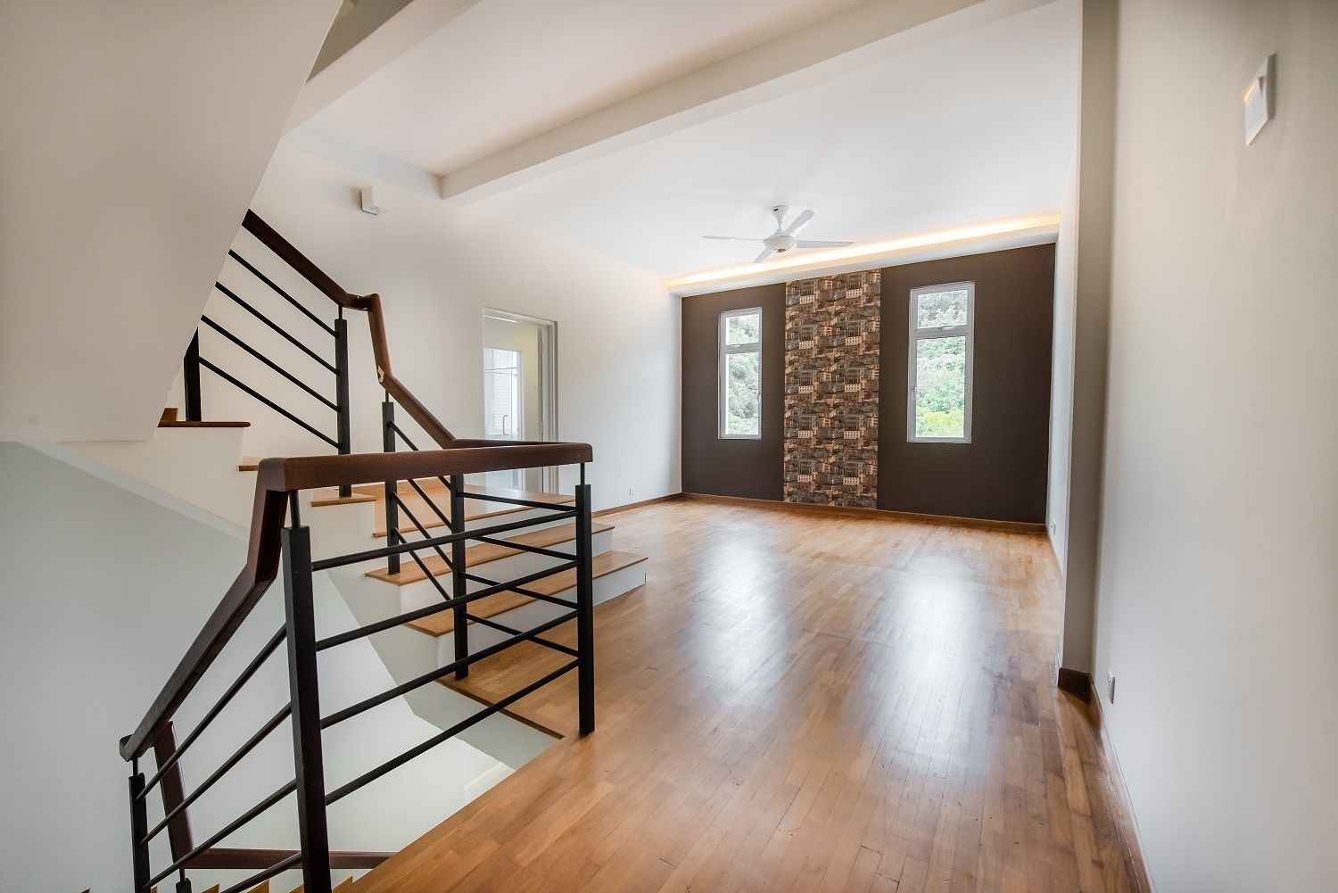 A spacious room with a wooden floor, a staircase with black railings, and a ceiling fan.