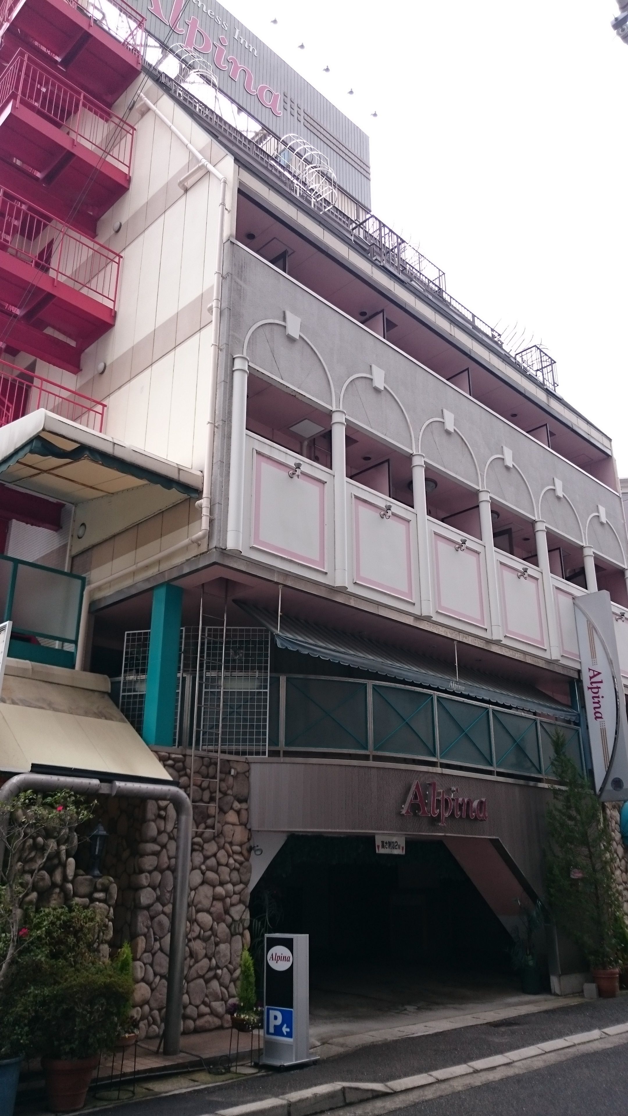 A multi-story building with a prominent 'Alpina' sign on the ground floor, a balcony with railings, and a parking sign.