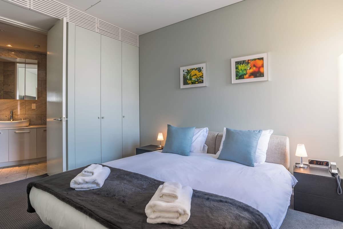 A hotel room with a bed, two pillows, a nightstand, a lamp, a clock, and a bathroom with a sink and mirror.