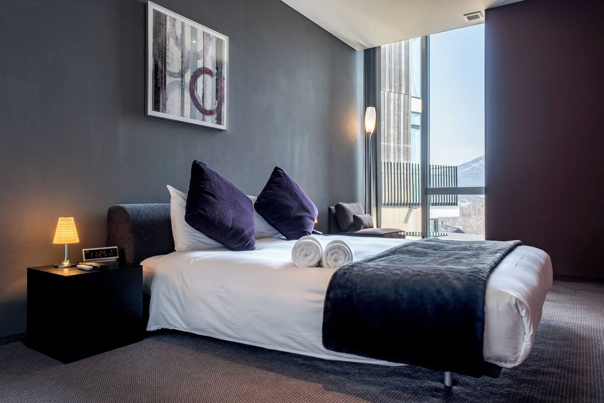 A modern hotel room with a large bed, a nightstand with a lamp, and a view of the city through large windows.